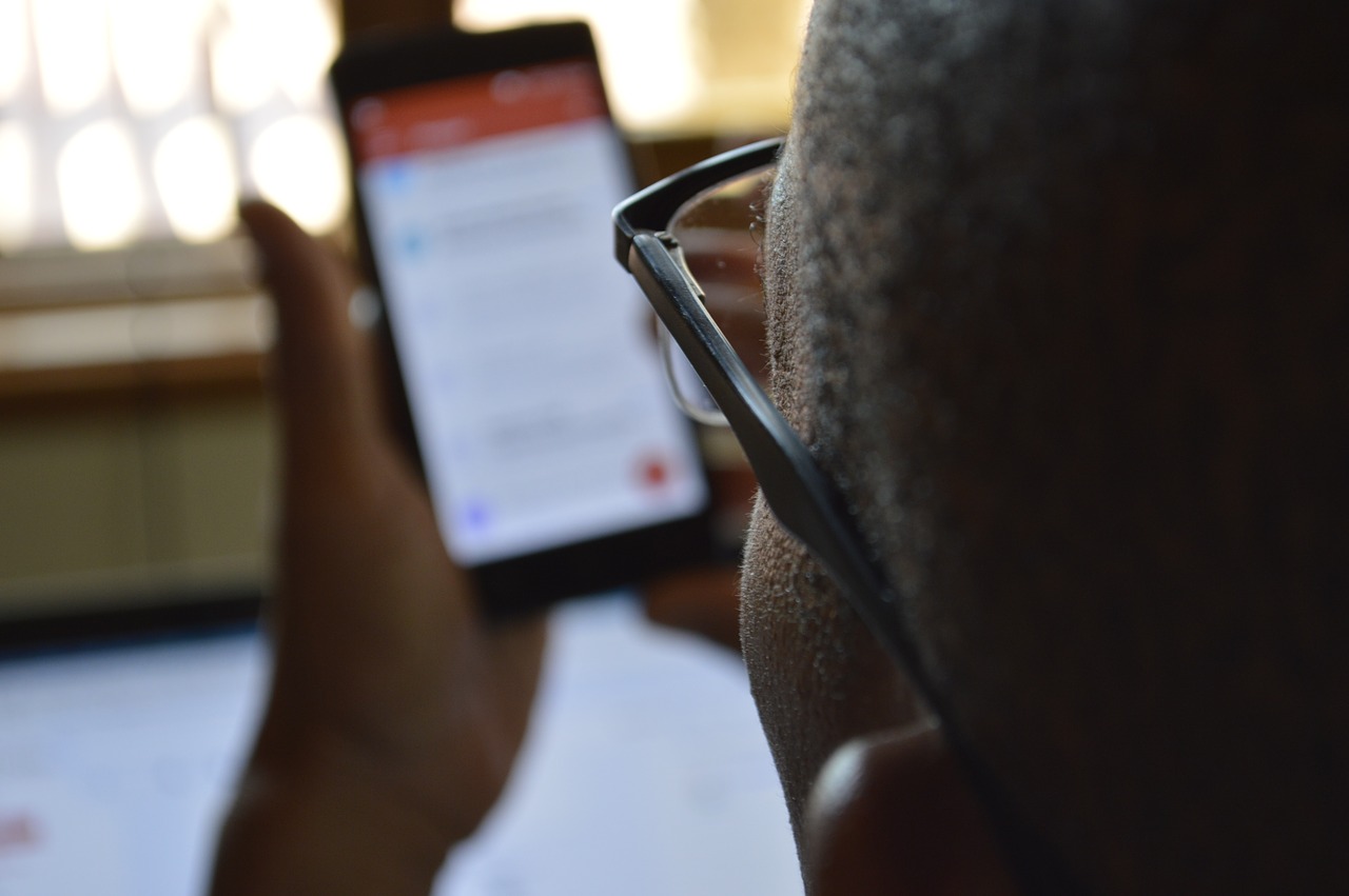 Foto de um homem negro segurando um celular nas mãos. Ele é negro, usa óculos e tem a cabeça raspada. 