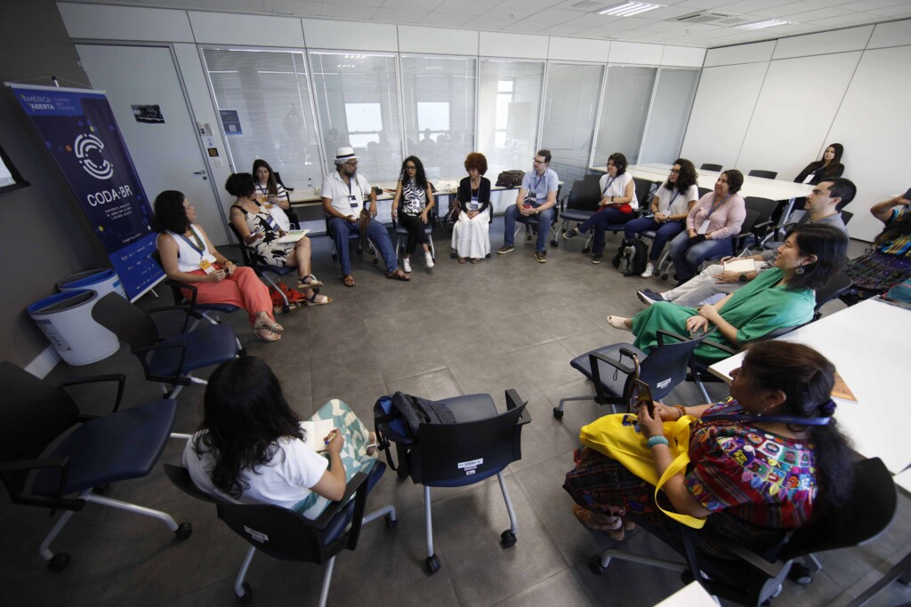 Pessoas sentadas em roda em cadeiras de escritório conversam dentro de uma sala, com um banner escrito Coda.Br no canto esquerdo