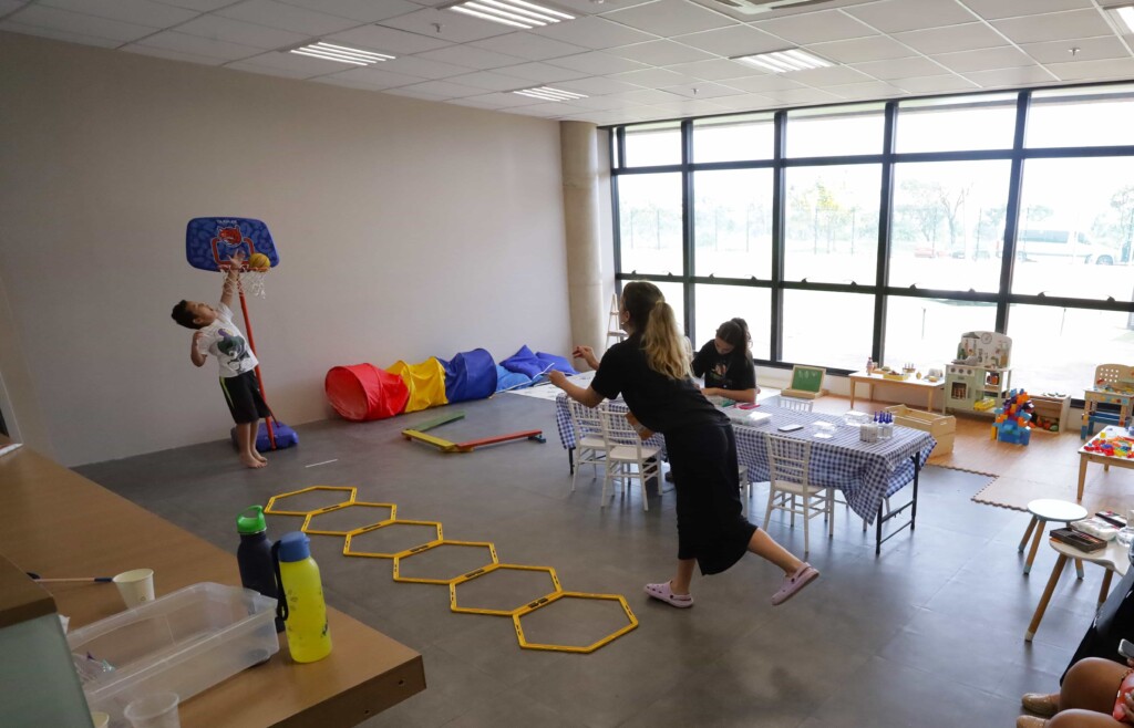 Mulher brincando de basquete com menino em uma sala interna, com argolas amarelas no chão e outros mobiliários