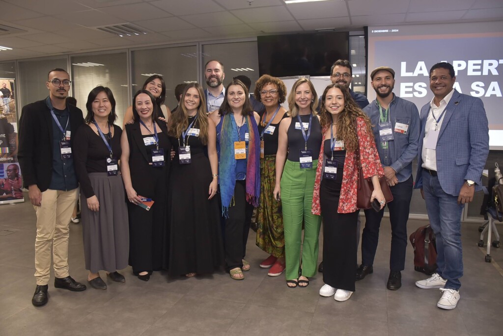 Treze pessoas com crachás posam juntas para foto em uma sala com uma projeção atrás